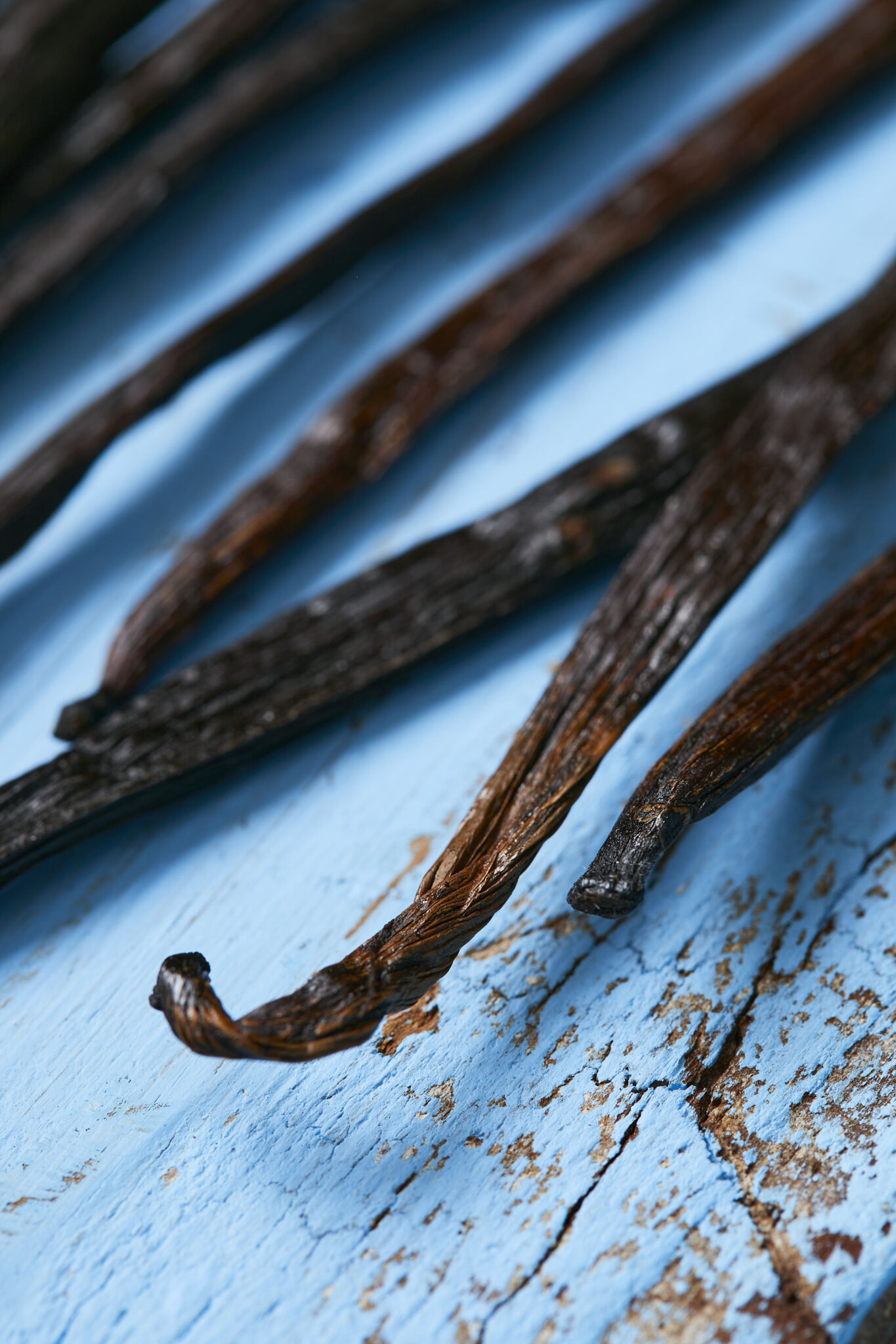 Vanilla beans are on a blue wooden surface. The bean pods are in a long, slender shape, have a dark blackish-brown color and a glossy appearance. 