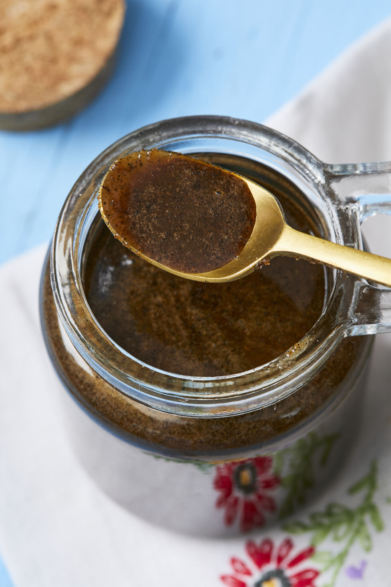 Homemade Vanilla Bean Paste is in a glass jar with a golden spoon holding some of it on top. The vanilla bean paste is in a dark blackish-brown color and a syrup consistency with speckles of vanilla seeds. 