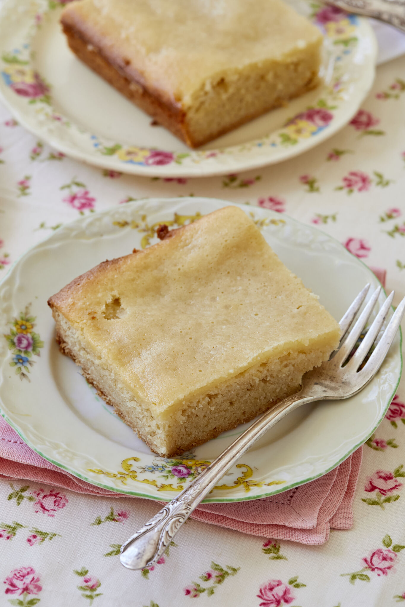 Two pieces of maple gooey butter cake on white plates with silver forks, on pink linen napkins on a floral tablecloth.