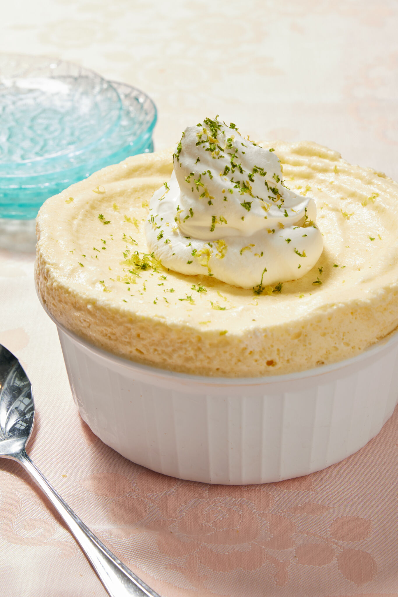 The silky smooth soufflé is perfectly set in a white ramekin, topped with a dollop of fluffy whipped cream and sprinkled with refreshing lemon and lime zest to boost the flavor. A silver serving spoon and a stack of light-blue glass dessert plates are on the left. 