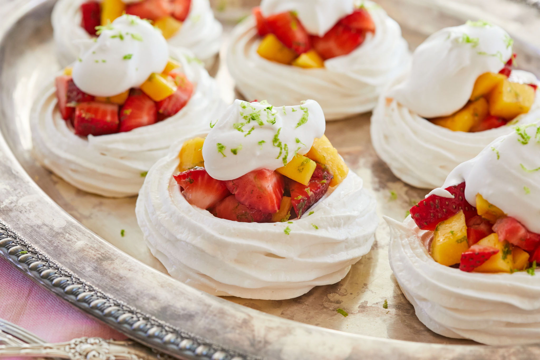 Heavenly Meringue Nests are placed on a silver platter. They are white layers of crispy billowy meringue loaded with irresistible tropical red strawberries and vibrant yellow mangoes, and adorned with velvety coconut cream, and green refreshing lime zest.