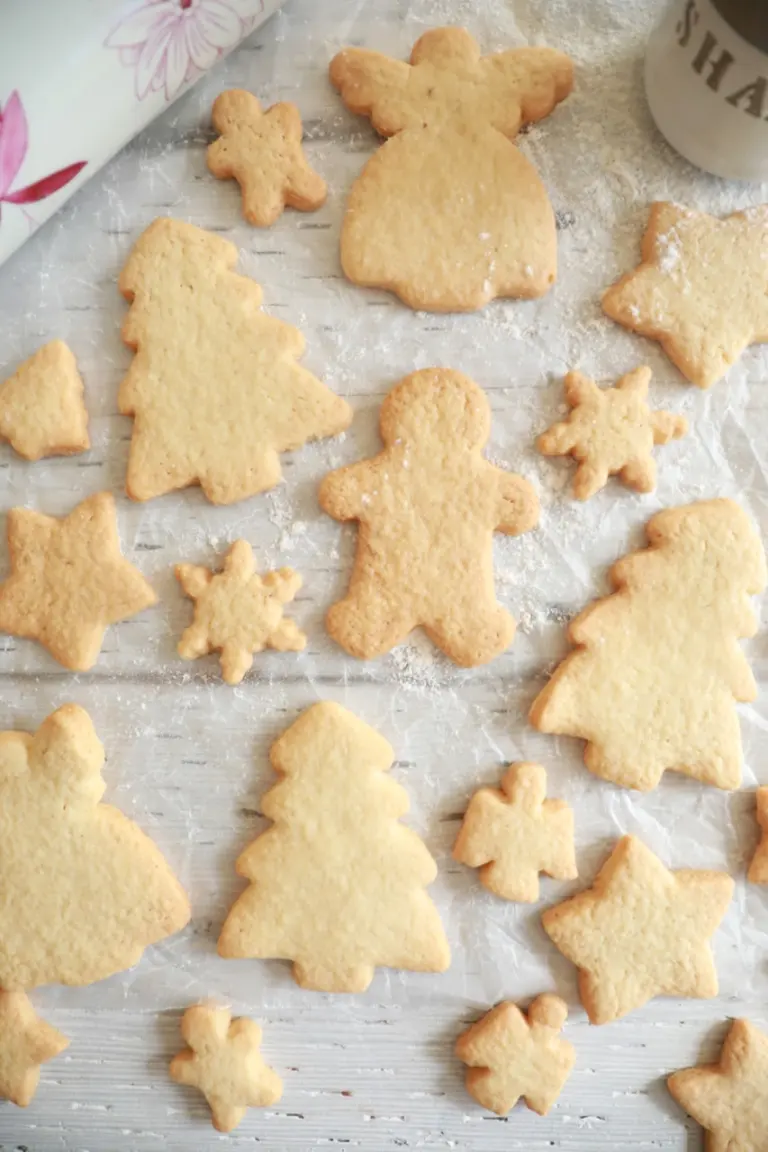 Homemade sugar cookies are presented on parchment paper. The sugar cookies are cut into different shapes, including a Christmas tree, an angel, snowflakes, and gingerbread men.